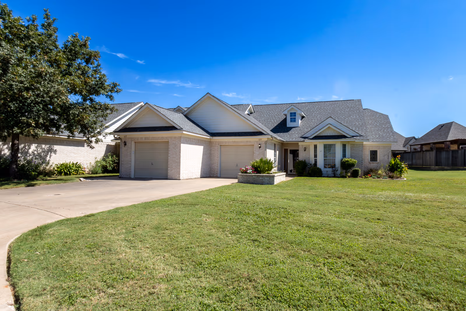 A single-story residential building with a light brick exterior, a gray shingled roof, and two garage doors. The house is surrounded by a well-maintained green lawn with a tree on the left side and some shrubs and flowers near the entrance. The sky is clear and blue.