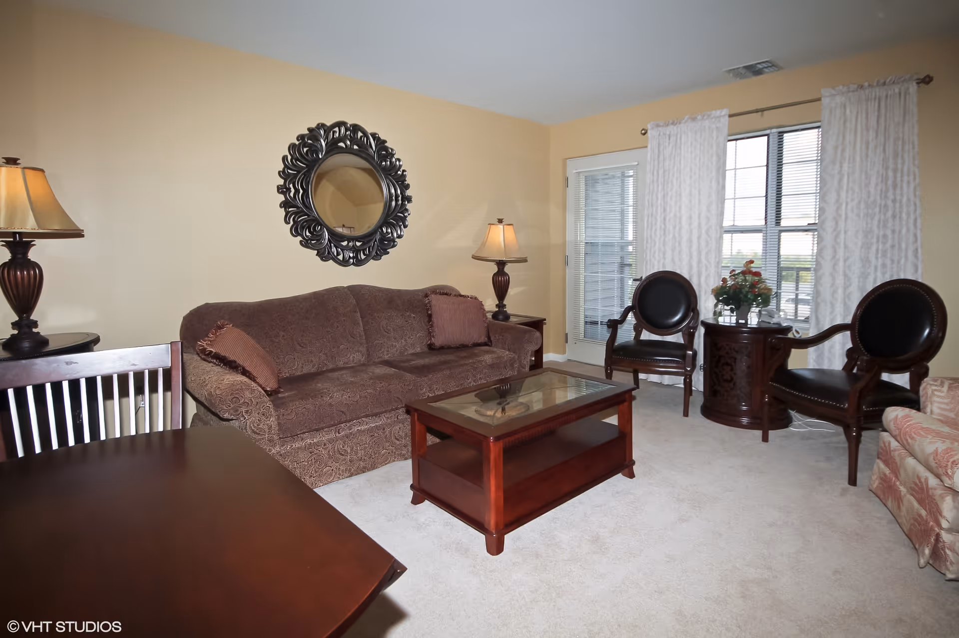 A cozy living room with a brown patterned sofa, two dark wooden chairs with black cushions, a glass-top wooden coffee table, and a round wooden side table with a flower arrangement. There are two table lamps with beige shades on either side of the sofa, a decorative round mirror on the wall, and a window with white curtains letting in natural light.