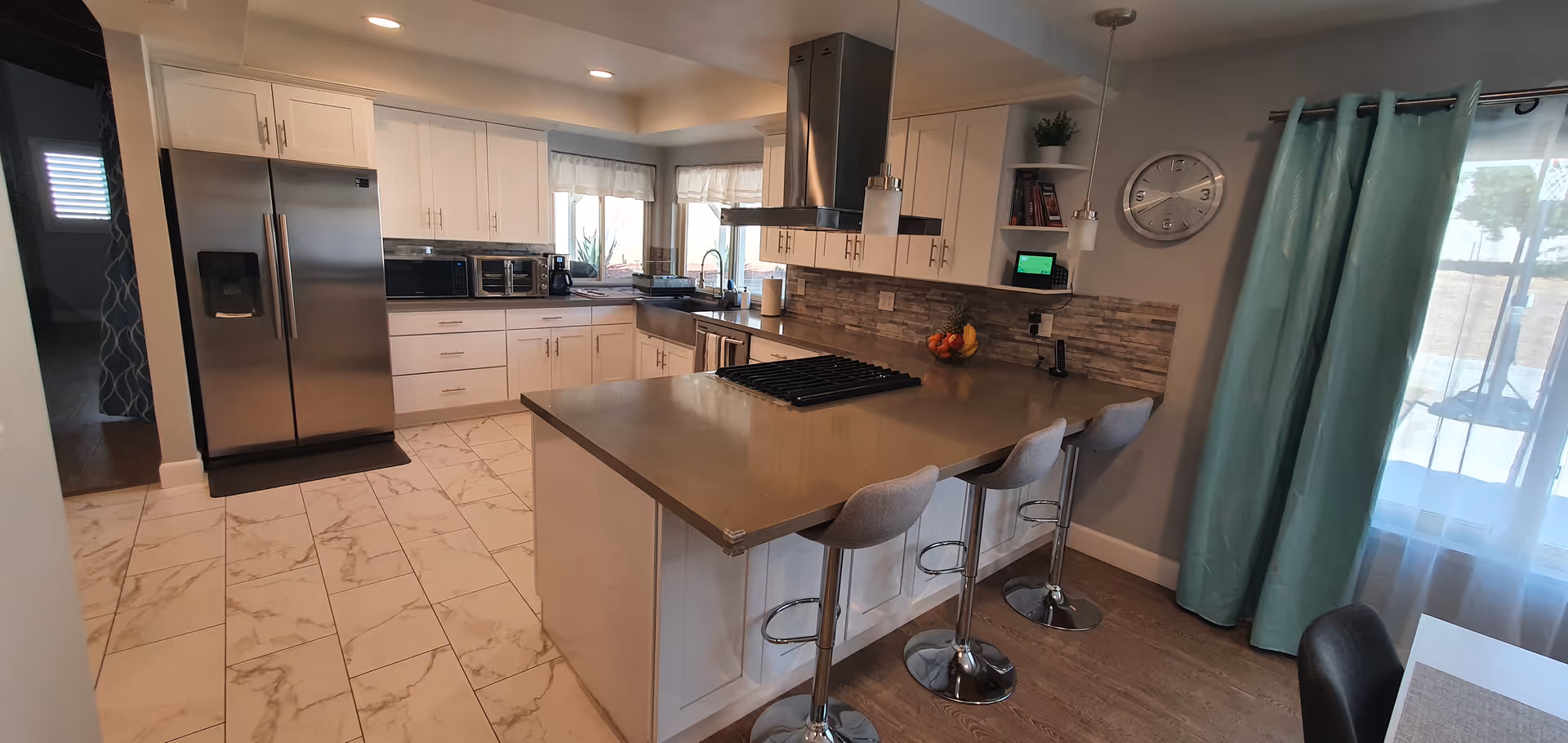 Modern kitchen with white cabinets, stainless steel refrigerator, built-in oven, microwave, and a large island with a cooktop and three bar stools. The kitchen has tiled flooring, a stone backsplash, and a window with a white valance. There is a wall clock and shelves on the right side, and a sliding glass door with teal curtains leading outside.