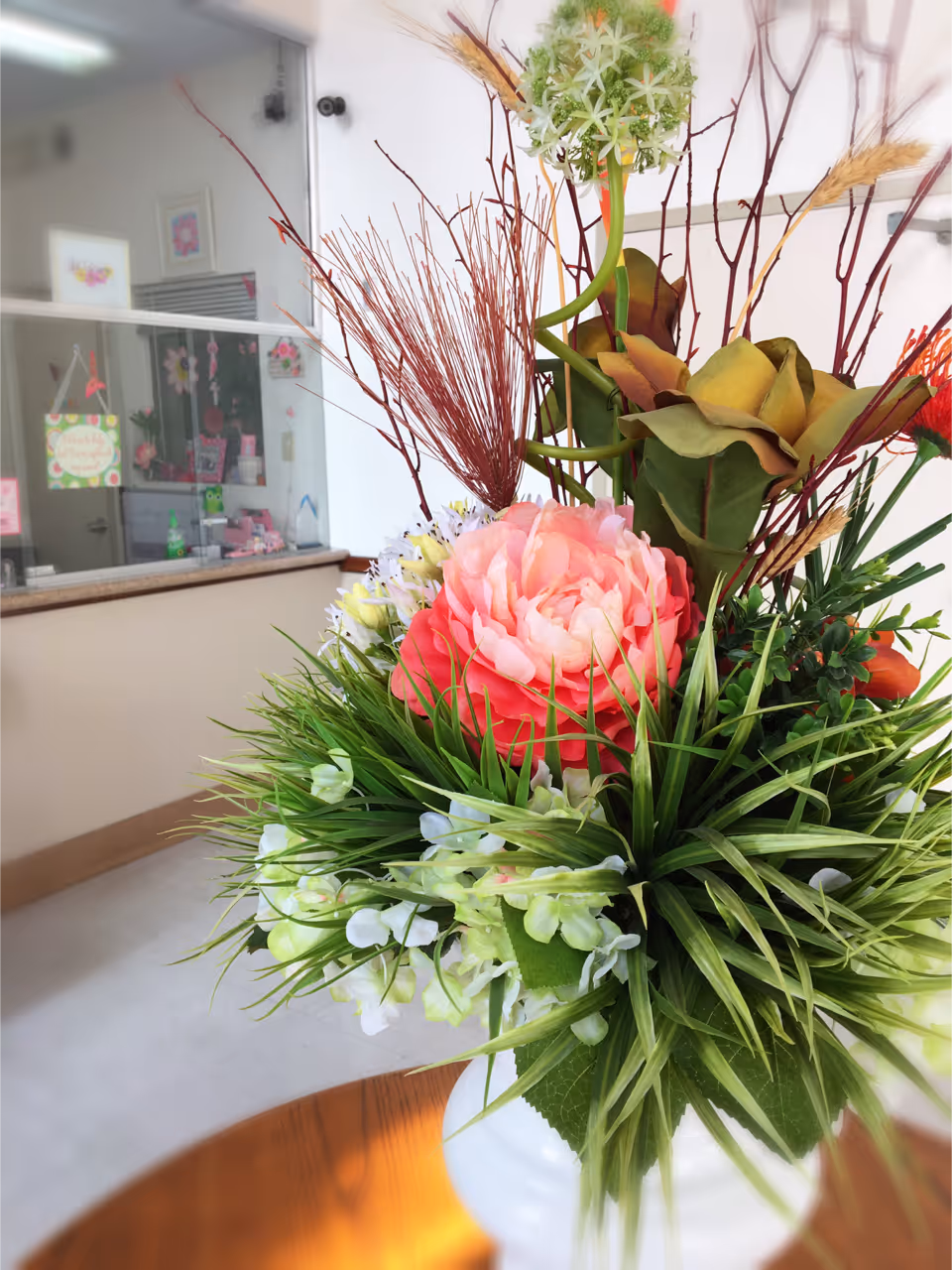 A close-up view of a floral arrangement with pink, white, and green flowers and various green leaves placed on a wooden table. In the background, there is a reception area with a glass partition and some decorations on the wall.