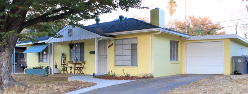 Single-story yellow house with a dark roof, a front porch with chairs and a small table, a large tree in the front yard, and an attached garage. The driveway and walkway lead up to the house, and there are some plants near the porch.