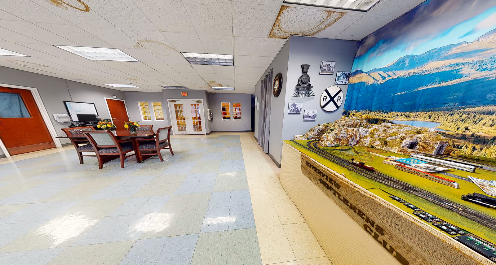 A spacious interior common room featuring a large model train display on the right and a table with chairs in the center.