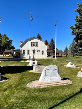 A white two-story building with a gabled roof and several windows, situated on a well-maintained lawn with multiple flagpoles and memorial stones. The sky is clear and blue, and there are tall evergreen trees in the background.