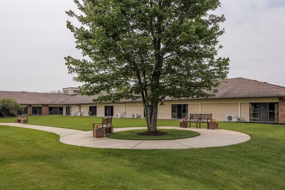 Circular paved courtyard with benches and a large tree in front of a single-story retirement facility building.