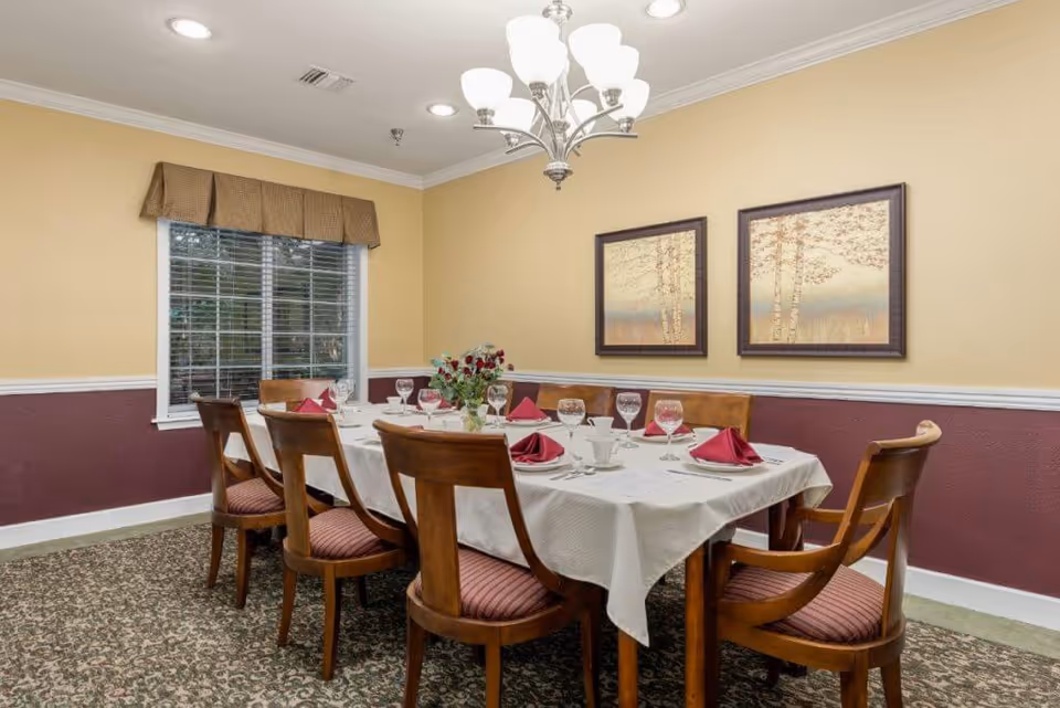 A dining room with a rectangular table set for eight people. The table is covered with a white tablecloth and has red folded napkins, white plates, cups, and wine glasses. There is a floral centerpiece in the middle of the table. The room has beige and maroon walls, a window with blinds and a valance, two framed pictures on the wall, and a chandelier hanging from the ceiling.