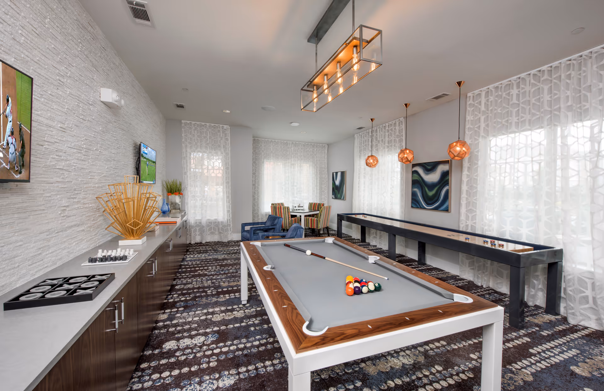 A modern game room with a pool table in the center, a shuffleboard table along the right wall, and a long cabinet on the left with chess and tic-tac-toe games on top. The room has patterned carpet, sheer white curtains on large windows, two blue armchairs, a small table with colorful chairs in the back, and two wall-mounted TVs showing sports. Pendant lights hang from the ceiling and abstract artwork decorates the walls.