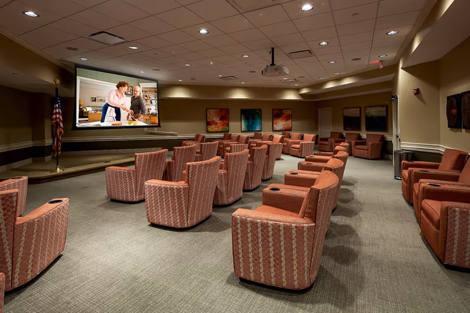 A spacious media room with multiple rows of cushioned armchairs facing a large screen showing two people cooking. The room has beige walls, ceiling lights, colorful abstract paintings on the walls, and an American flag near the screen.