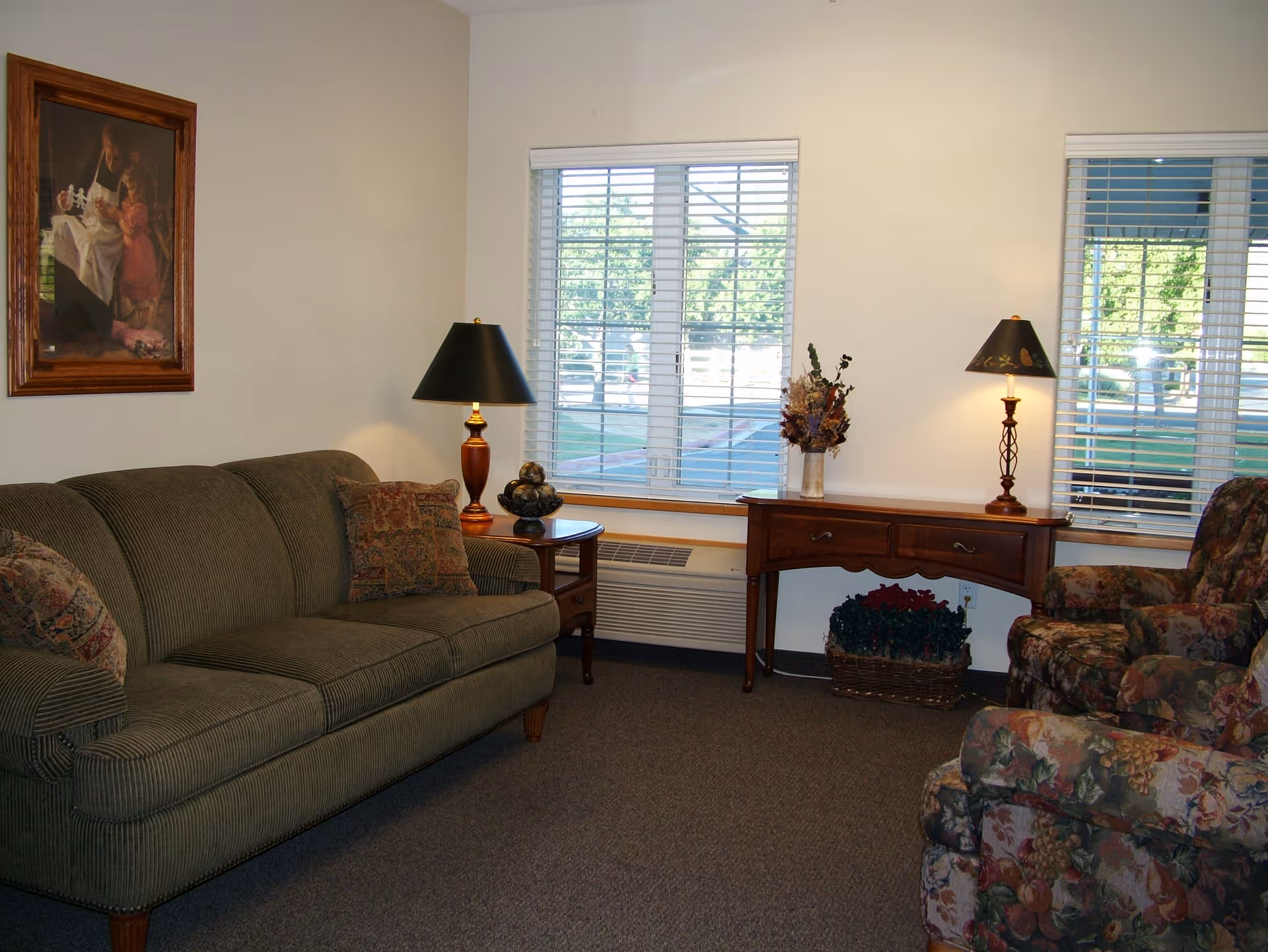 Cozy living room with a green sofa, floral armchairs, wooden side table and lamps beneath two windows with blinds.