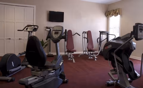 A small fitness room with various exercise equipment including stationary bikes, weight machines, and a treadmill. The room has beige walls, a red carpet, a window with a valance, and a wall-mounted television.
