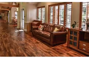 Interior view of a senior living facility hallway with wooden flooring, a brown leather sofa with decorative pillows, large windows with wooden blinds, and a wooden cabinet with drawers and a plant on top.