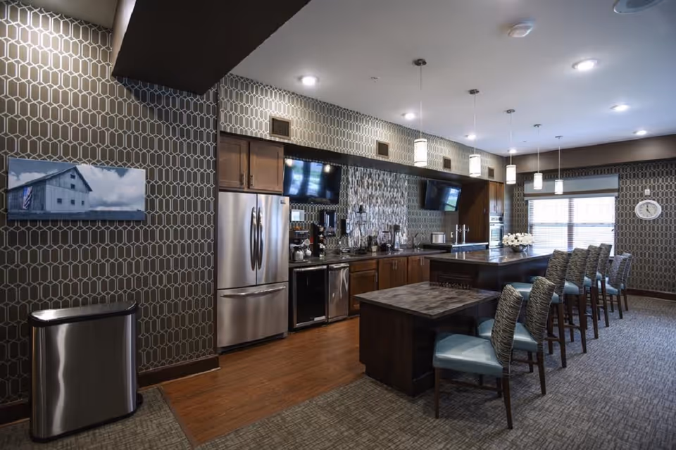 Communal kitchen and dining area with stainless steel appliances, a long island with pendant lights and barstools, and patterned wallpaper.