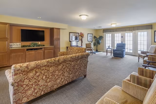 A spacious living room in a senior living facility with floral patterned sofas, armchairs, a large flat-screen TV mounted on a wooden cabinet, framed artwork on the walls, and large windows with glass doors allowing natural light to fill the room.