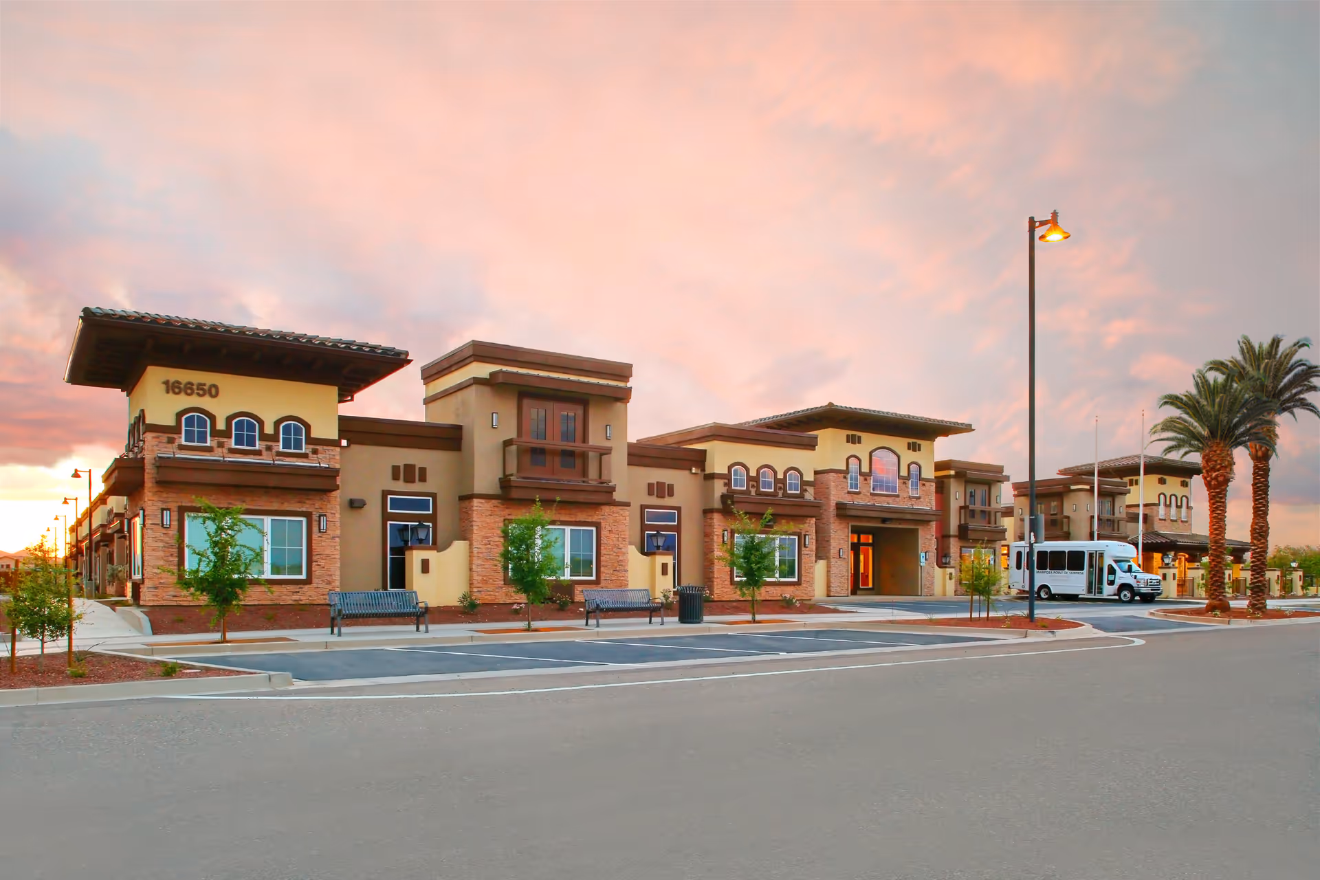 Exterior view of Vineyard Park of Surprise senior living facility at sunset, showing a modern building with stone and stucco facade, arched windows, benches, small trees, palm trees, a street lamp, and a white shuttle bus parked near the entrance.
