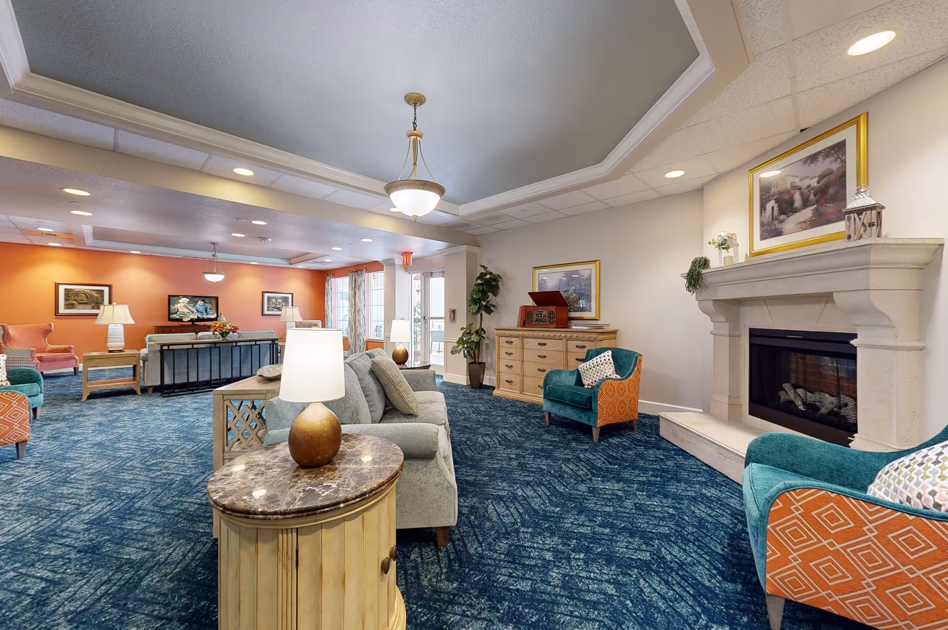 A spacious senior living facility lounge area with blue carpet, multiple seating options including sofas and armchairs with patterned cushions, a stone fireplace, framed artwork on the walls, and several table lamps providing warm lighting.