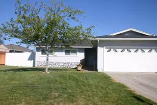 Single-story residential building with a two-car garage, manicured lawn, and tree in front.