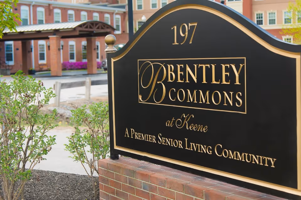 A black and gold sign reading '197 Bentley Commons at Keene A Premier Senior Living Community' with a brick building and greenery in the background.