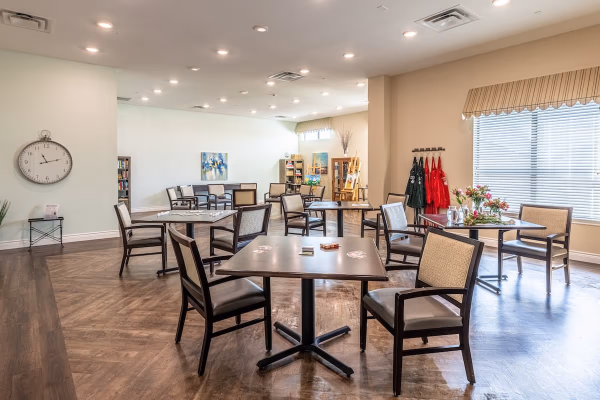 Spacious common dining/activity room with multiple tables and chairs, a large wall clock, bookshelf, and a window with blinds.