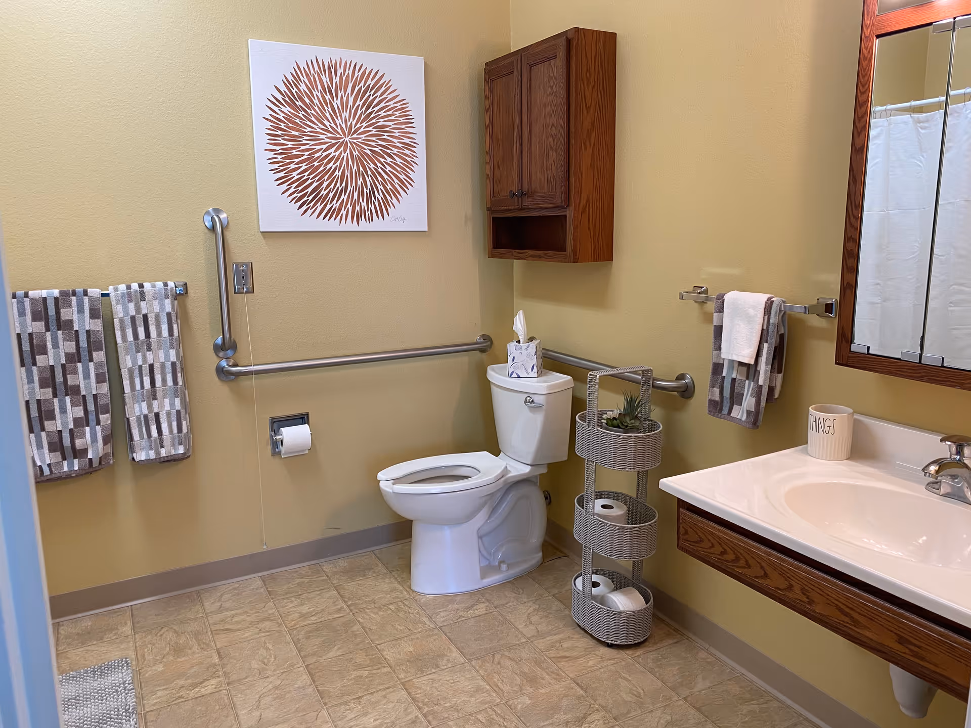 A clean and accessible bathroom with beige walls and tiled floor. The room features a white toilet with a tissue box on top, a wooden cabinet mounted on the wall above the toilet, and a three-tiered wicker storage unit holding toilet paper rolls and a small plant. There are grab bars installed around the toilet for accessibility. Two patterned towels hang on a towel rack to the left, and another towel hangs on a rack next to a white sink with a wooden vanity. A large mirror is mounted above the sink, and a decorative cup labeled 'THINGS' sits on the sink counter. A piece of wall art with a circular, spiky design in brown tones is hung above the grab bar on the left wall.