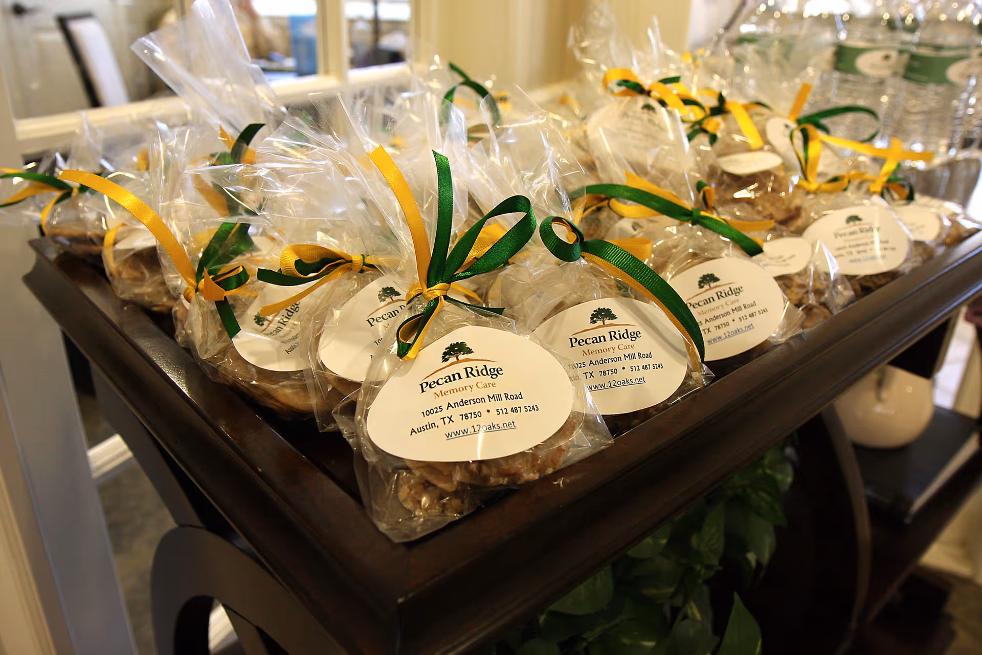 Several small clear plastic bags tied with green and yellow ribbons, each containing pecan treats with labels that read 'Pecan Ridge Memory Care, 10025 Anderson Mill Road, Austin, TX 78750, 512 487 5243, www.12oaks.net', displayed on a dark wooden table.