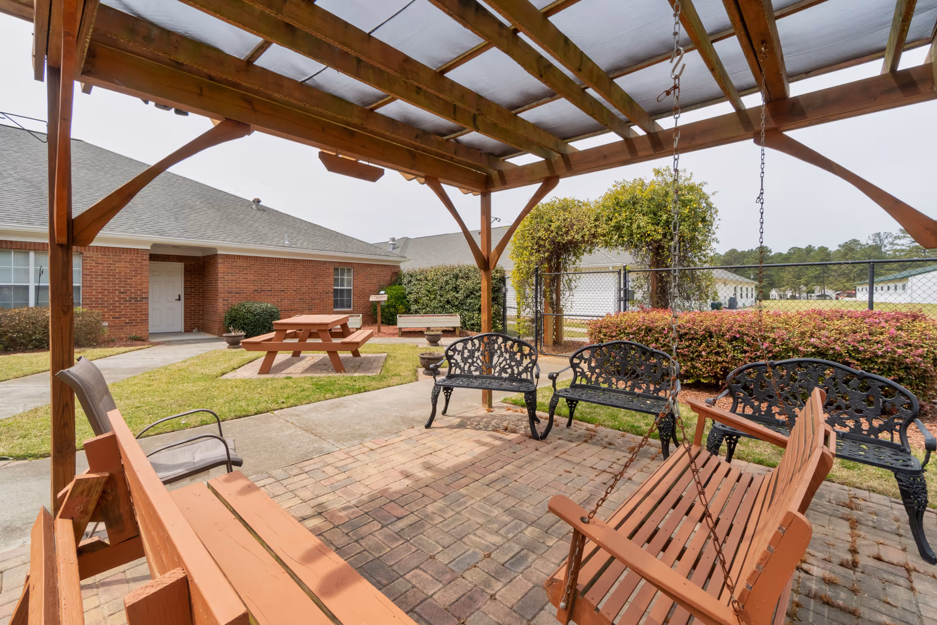Outdoor seating area at Merryvale Assisted Living featuring a wooden pergola with a translucent roof, a wooden swing bench hanging from the pergola, several metal benches with decorative backs, a picnic table on a grassy area, and a brick building in the background.
