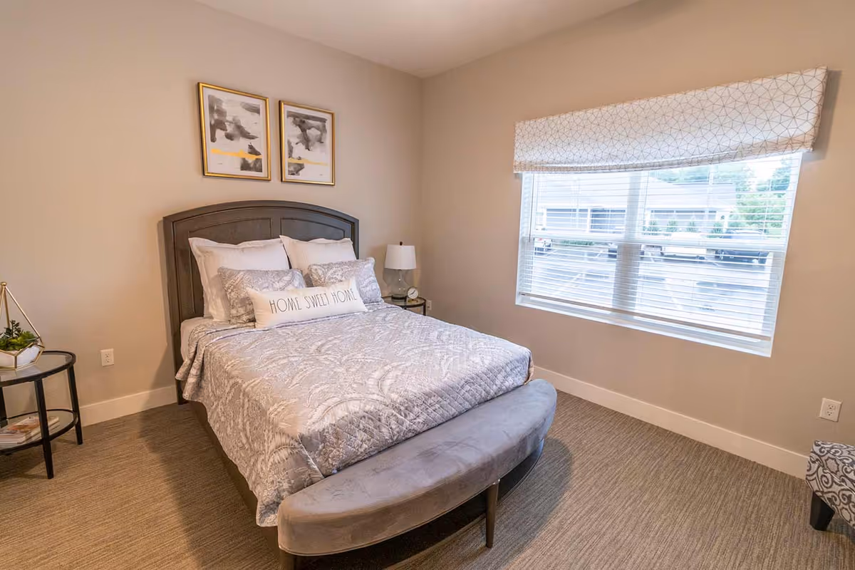 A cozy bedroom with a neatly made bed featuring a gray patterned comforter and multiple pillows, including one that says 'HOME SWEET HOME'. There is a cushioned bench at the foot of the bed, a small round side table with a plant, and a lamp on another side table near the window. The window has blinds and a patterned valance, and two framed abstract paintings hang on the wall above the bed.