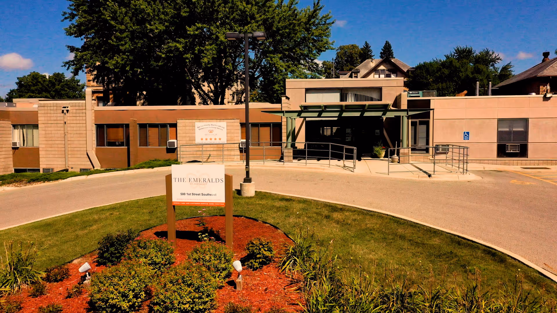 Exterior view of The Emeralds at Faribault senior living facility showing a single-story building with a ramp entrance, surrounded by greenery and a small landscaped area with bushes and a sign displaying the facility's name and address.