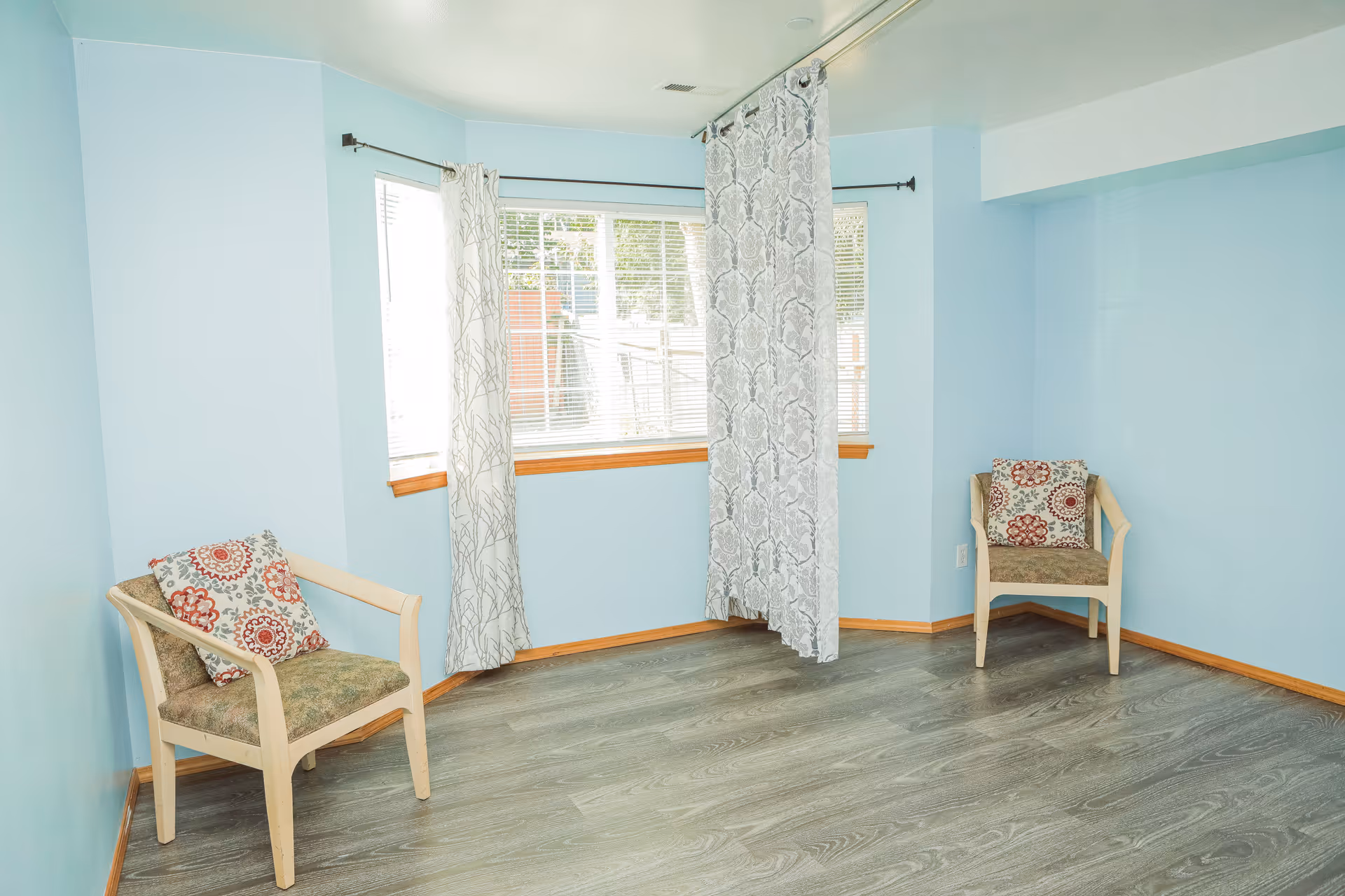 A small room with light blue walls and wood trim around the windows and floor. There are two wooden chairs with patterned cushions placed against the walls. The room has large windows with white blinds and two patterned curtains hanging from a black curtain rod. The floor is covered with gray wood-like laminate.