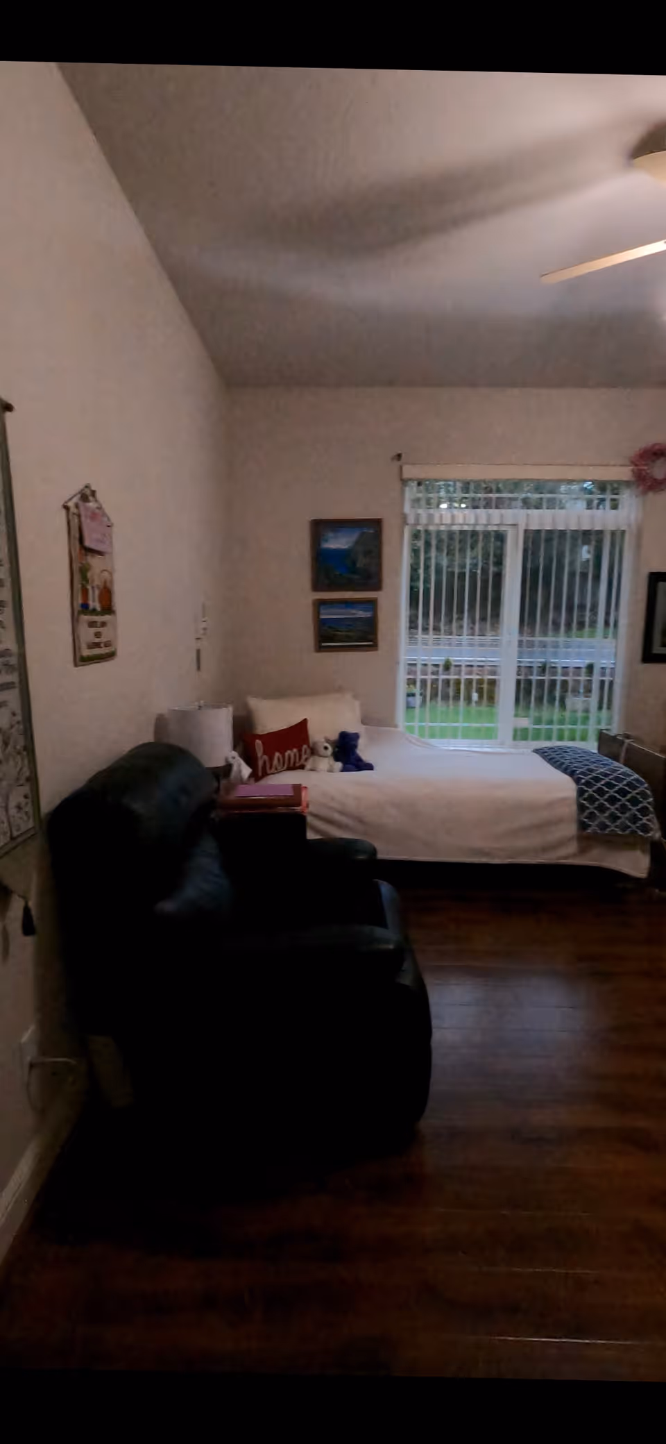 A cozy bedroom with a single bed against the far wall, decorated with pillows including one that says 'home' and a small stuffed animal. There is a large window with vertical blinds letting in natural light. A black recliner chair is positioned in the foreground on the left side. The room has wooden flooring and light-colored walls with framed pictures hanging.