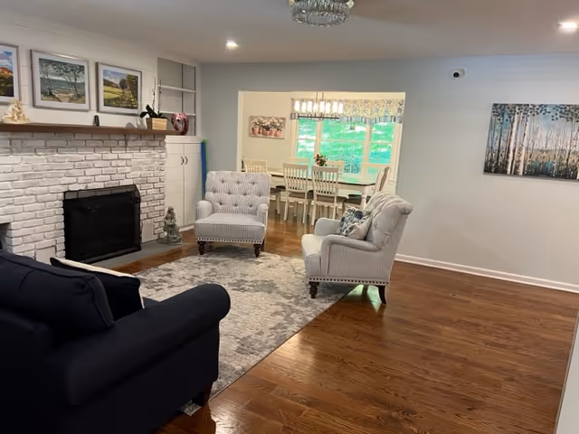 Spacious living room with a white brick fireplace, upholstered chairs and sofa around an area rug, and a dining table visible through a pass-through window.