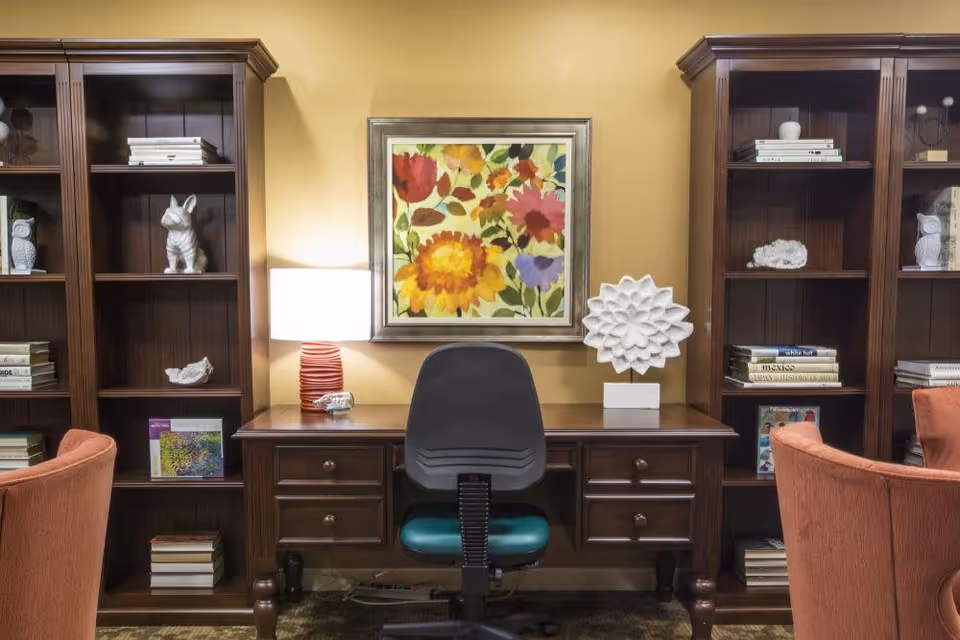 A cozy interior space featuring a dark wooden desk with drawers, a black office chair with a green seat cushion, and two tall wooden bookshelves on either side filled with books and decorative items. A colorful floral painting hangs on the wall above the desk, flanked by a table lamp with a red base and a white decorative sculpture. Two orange upholstered chairs are partially visible in the foreground.