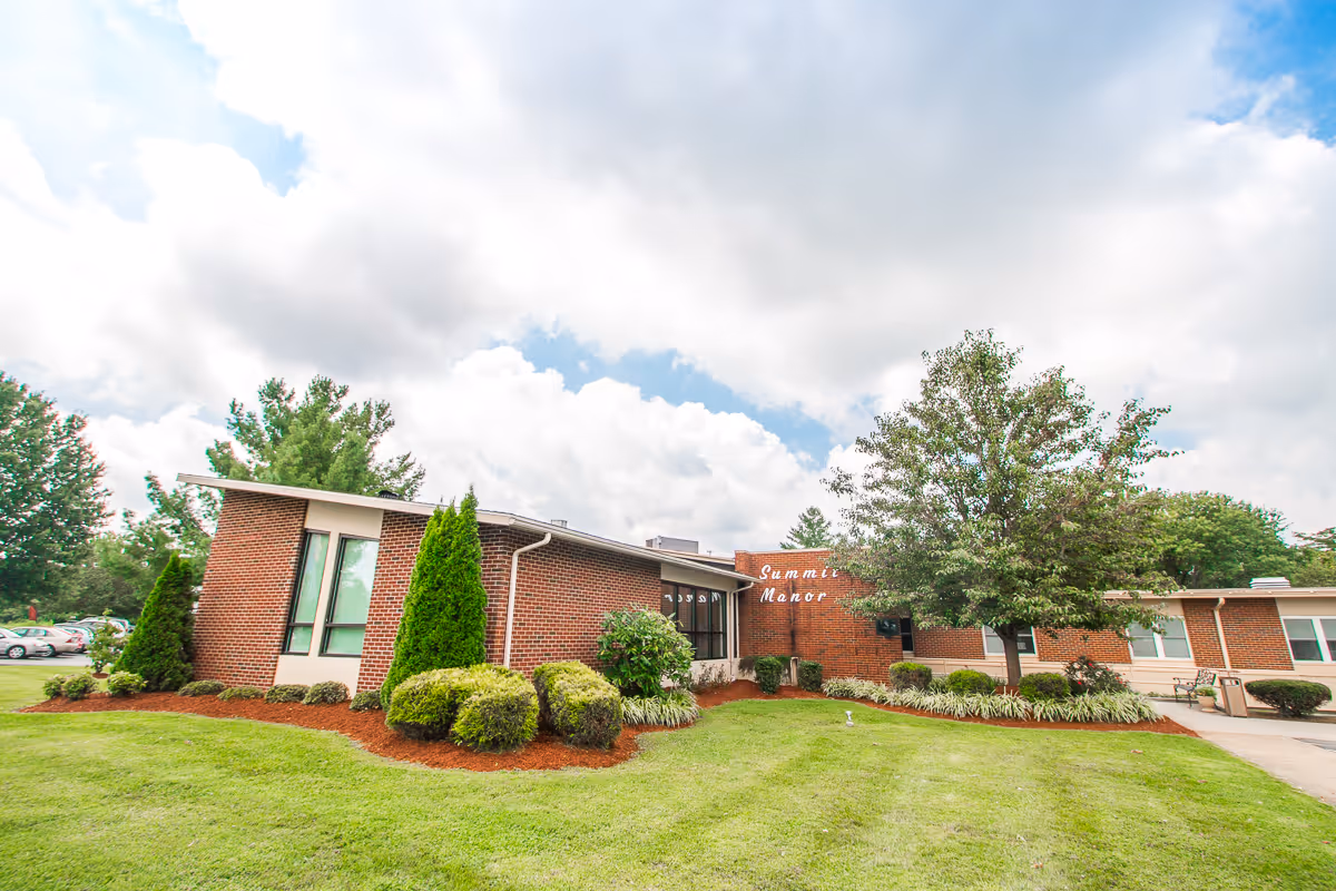 Exterior view of a single-story brick building with large windows, surrounded by green grass, bushes, and trees under a partly cloudy sky. The building has a sign that reads 'Summit Manor'.