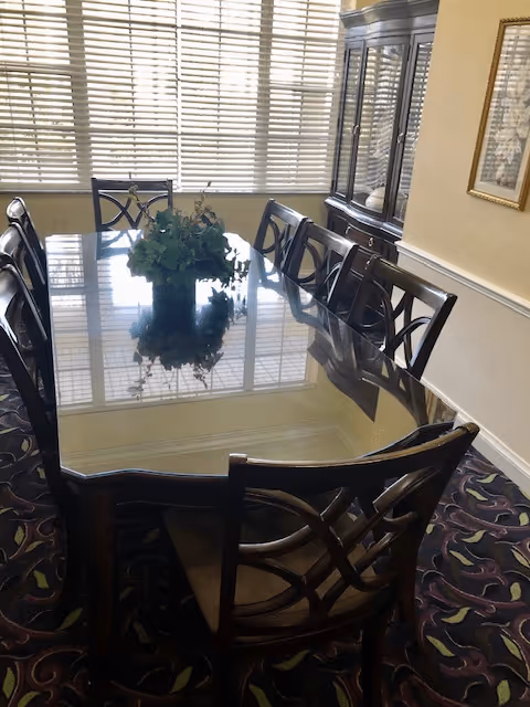A dining room with a large rectangular glass-top table surrounded by eight wooden chairs with cushioned seats. A green plant centerpiece sits on the table. Behind the table is a large window with blinds, and to the right is a wooden china cabinet with glass doors. The room has patterned carpet and a framed picture on the wall.