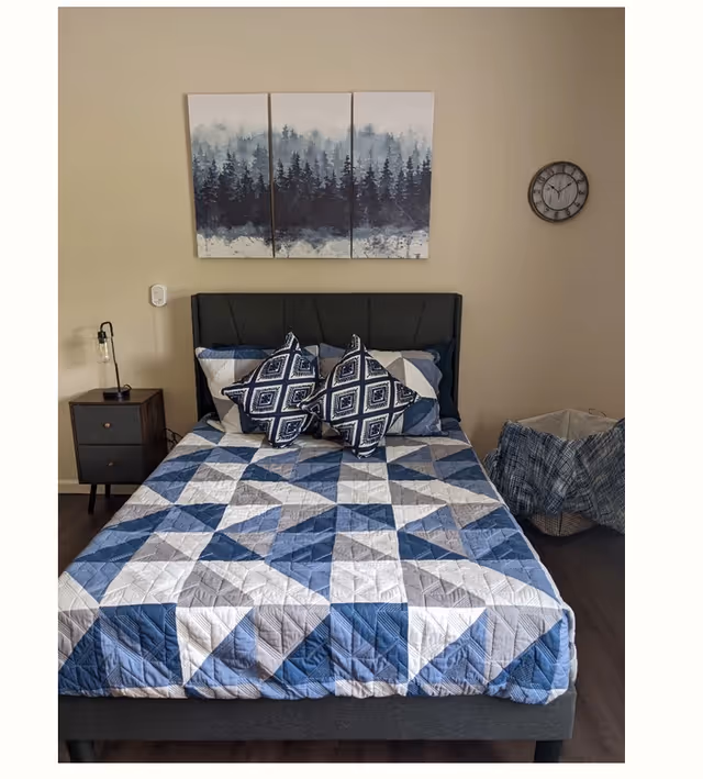 A neatly made bed with a blue, white, and gray geometric patterned quilt and two decorative pillows with a diamond pattern. Above the bed is a three-panel wall art depicting a misty forest. To the left of the bed is a small dark wooden nightstand with a modern lamp. On the right side, there is a basket covered with a blue plaid cloth. A round wall clock is mounted on the beige wall above the basket.