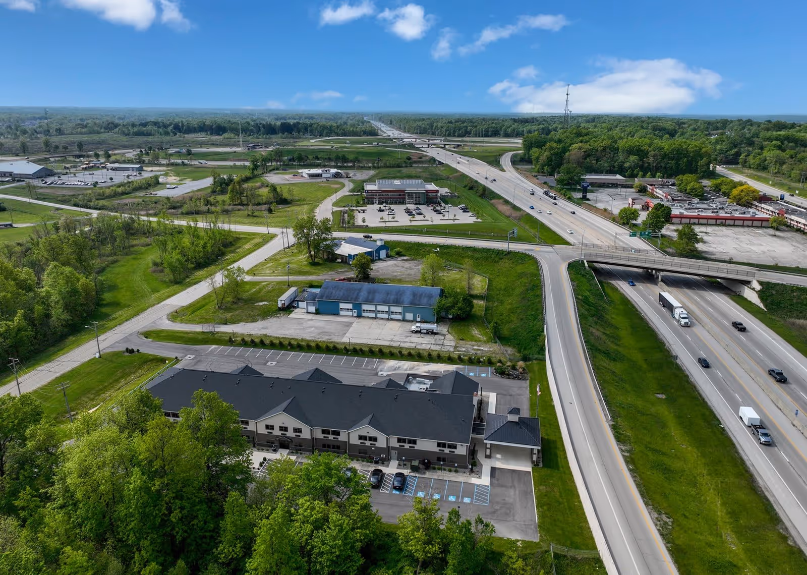 Aerial view of a senior living facility named Hines Hill Senior Living surrounded by green trees and grass. The building is located near a highway with multiple lanes and an overpass. Several parking lots and other buildings are visible in the surrounding area under a partly cloudy blue sky.