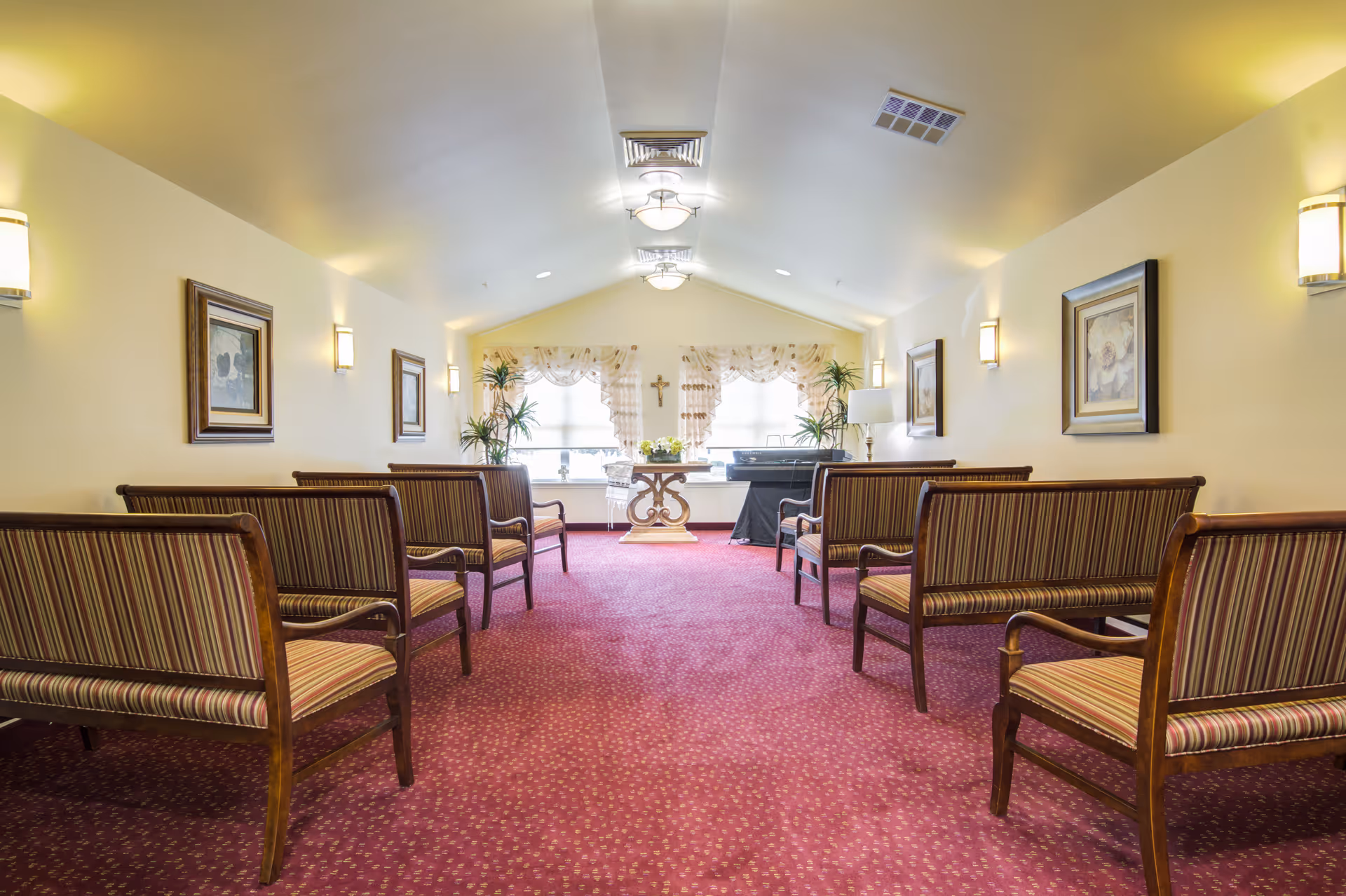 A small chapel or meditation room with rows of wooden benches with striped cushions facing a table with flowers and a cross on the wall. The room has a red carpet, cream-colored walls, framed floral artwork, wall sconces, and windows with sheer curtains letting in natural light.