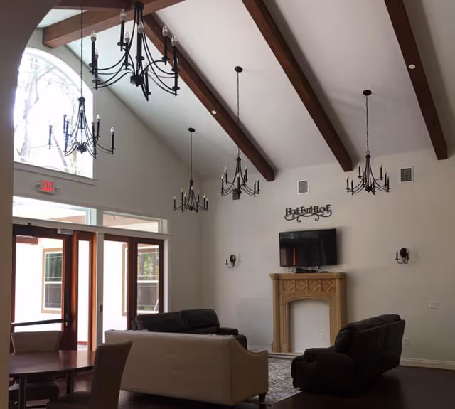 A spacious living room with high vaulted ceilings featuring exposed wooden beams and multiple black chandeliers. The room has large windows and glass doors letting in natural light. There are several sofas arranged around a decorative fireplace with a mounted flat-screen TV above it. The walls are painted white, and the floor is dark wood.