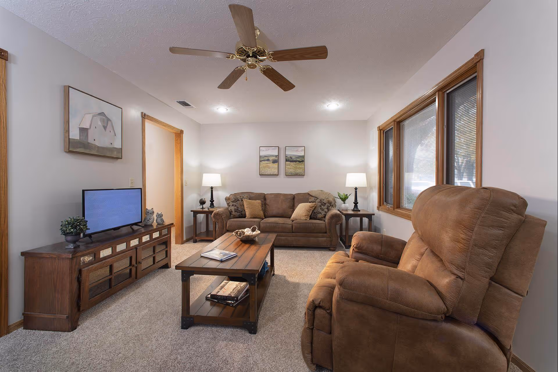 A cozy living room with a brown sofa and matching recliner chair, a wooden coffee table with books and decorative items, a wooden TV stand with a flat-screen TV and small plants, two side tables with lamps, and three windows with blinds. The walls are light-colored with framed artwork, and a ceiling fan with wooden blades is mounted on the ceiling.
