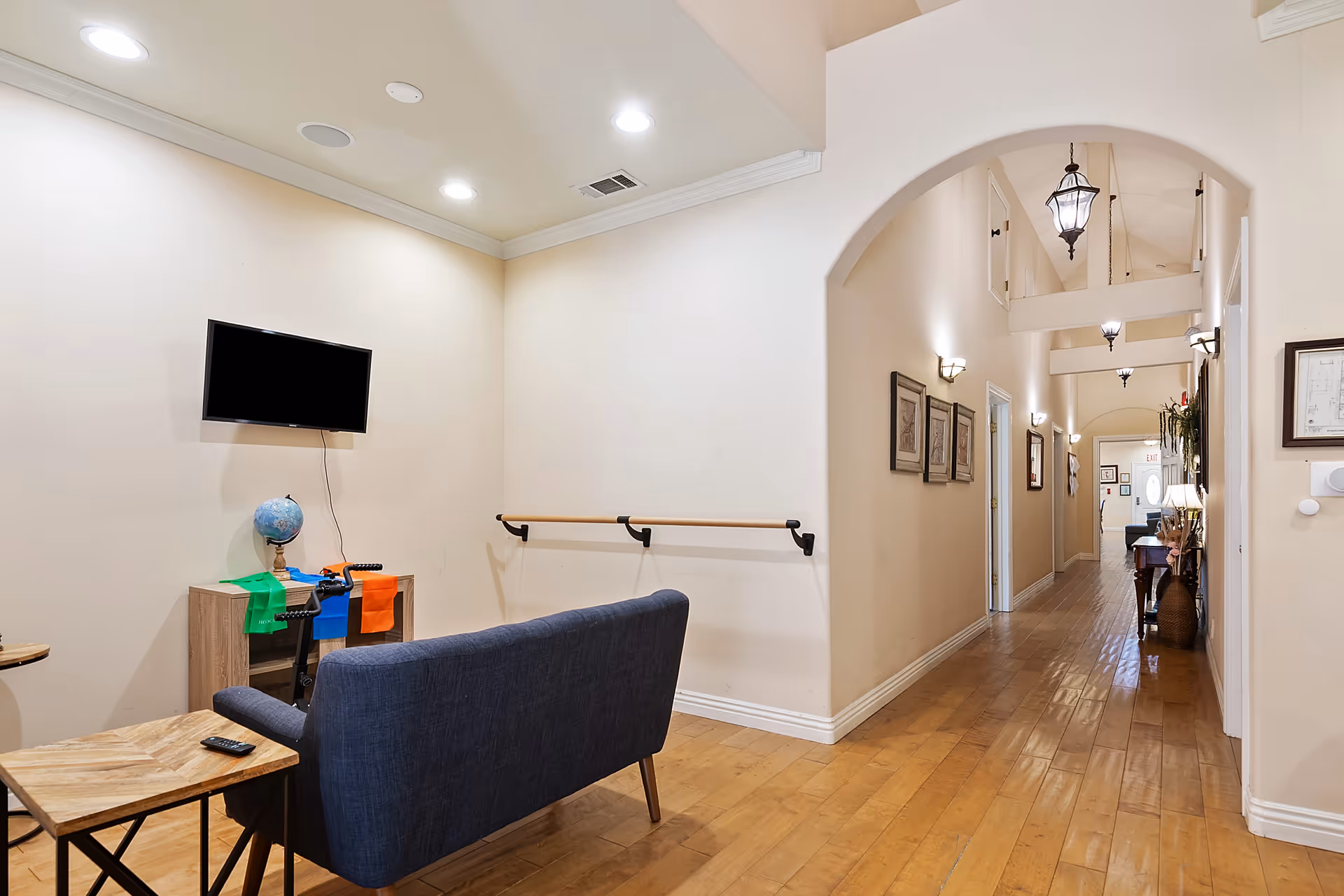 Interior view of a senior living facility hallway with wooden flooring and beige walls. On the left side, there is a small seating area with a dark blue couch facing a wall-mounted flat-screen TV above a small wooden cabinet with a globe and colorful resistance bands. The hallway features arched doorways, framed pictures on the walls, and hanging lantern-style light fixtures on the ceiling.