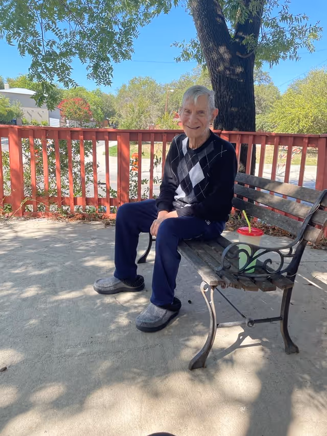 An elderly man wearing a black and white argyle sweater and navy pants sitting on a wooden bench with metal armrests outdoors. There is a red fence and green trees in the background, and a green drink with a red lid and straw placed on the bench beside him. The scene is shaded by tree branches overhead.