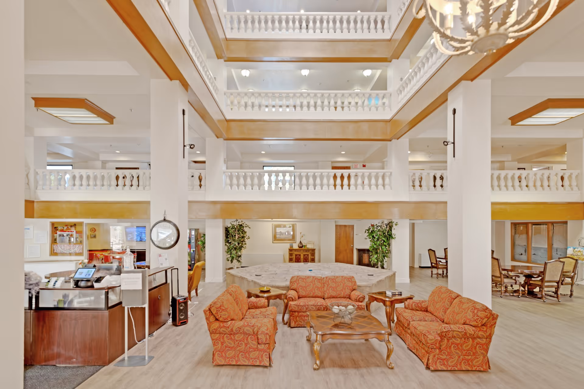 Spacious and well-lit senior living facility common area with three patterned orange sofas arranged around a wooden coffee table. The room features white pillars, a high ceiling with multiple balconies, and decorative railings. There are plants and framed artwork on the walls, and additional seating areas with tables and chairs are visible in the background.