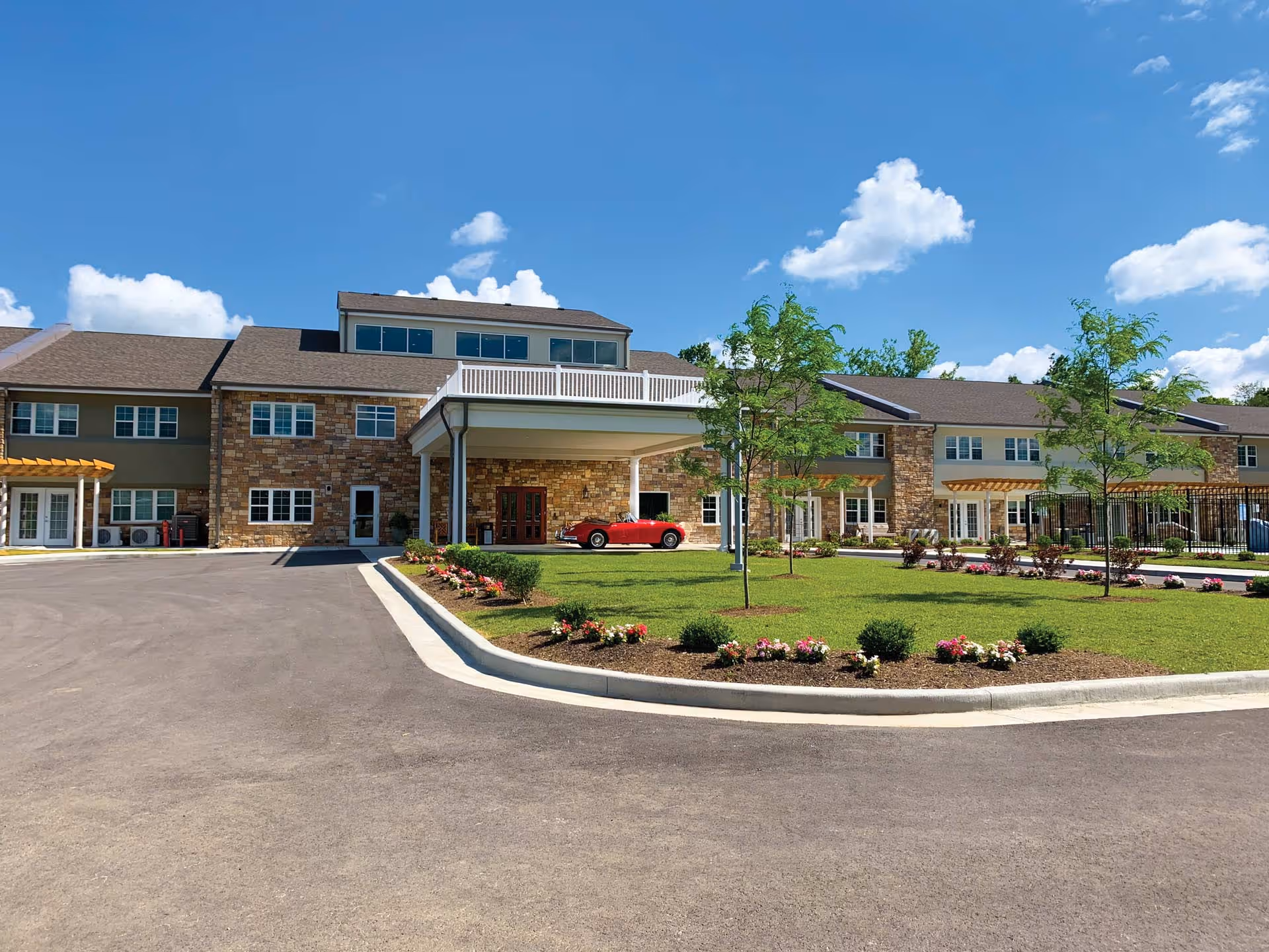 Front exterior of a senior living building with a covered entrance, circular driveway, landscaped lawn and a red car parked under the portico.