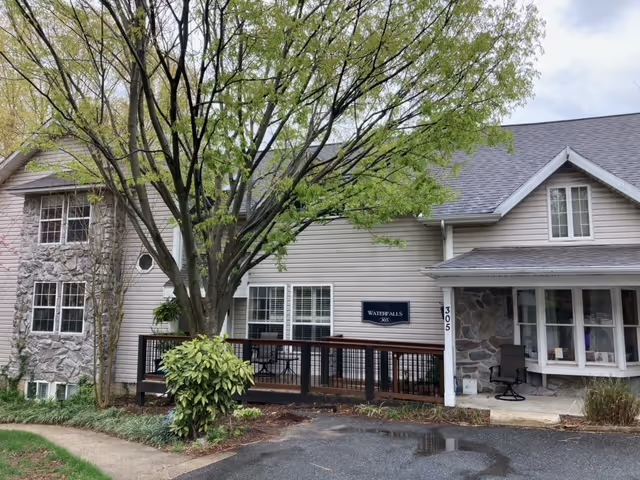 Front exterior of a two-story memory care home with a wooden deck, large tree, and a 'Waterfalls' sign by the entrance.