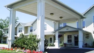 Exterior view of Parkwood Meadows Assisted Living facility showing a covered entrance with white pillars, a driveway, and well-maintained landscaping including bushes and flowers.