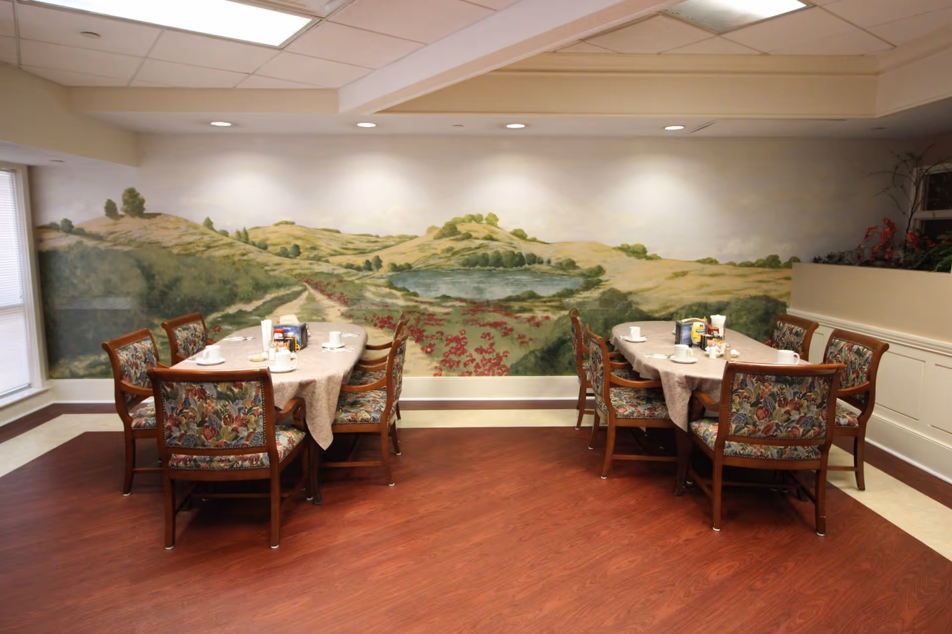 Small dining area with two tables and upholstered chairs set before a large painted landscape mural.