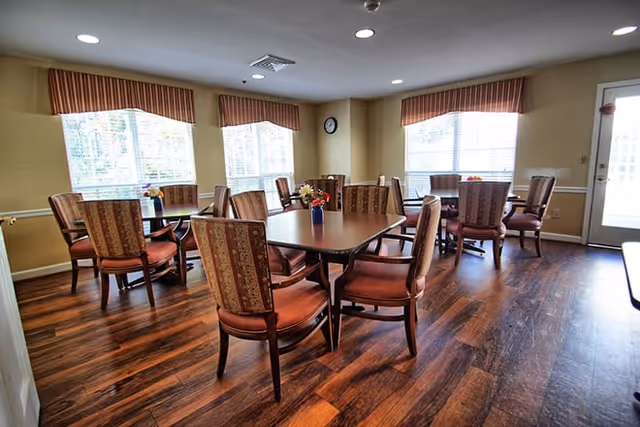 A bright dining room with several wooden tables and cushioned chairs arranged neatly. The room has large windows with striped valances allowing natural light to fill the space. There is a clock on the wall and a door with a window leading outside. The floor is made of dark wood.