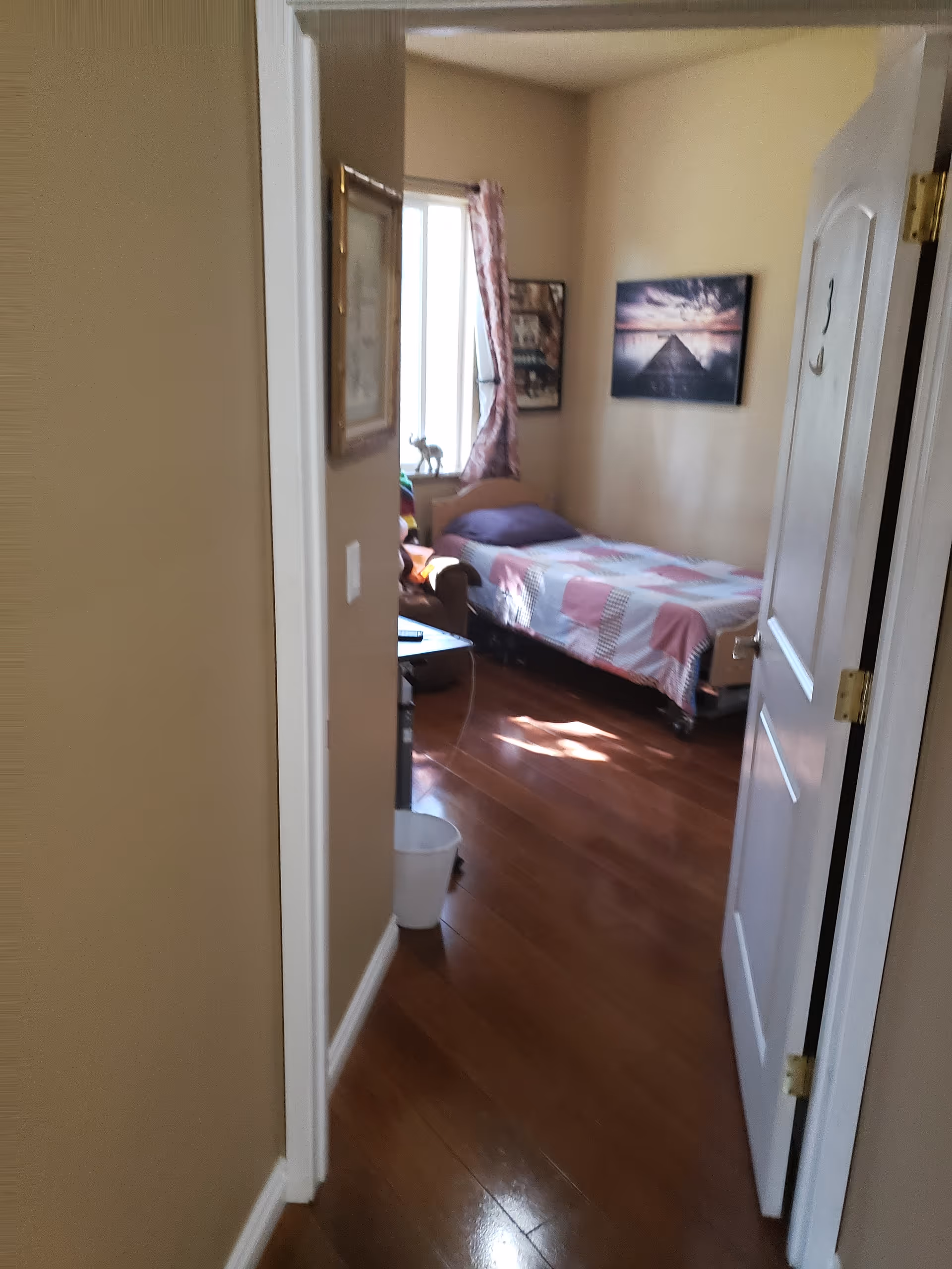 View through an open door into a small bedroom with a single bed covered with a pink and white patchwork quilt. The room has wooden flooring, a window with pink curtains, a framed picture on the wall, and a small table with a chair next to the bed.