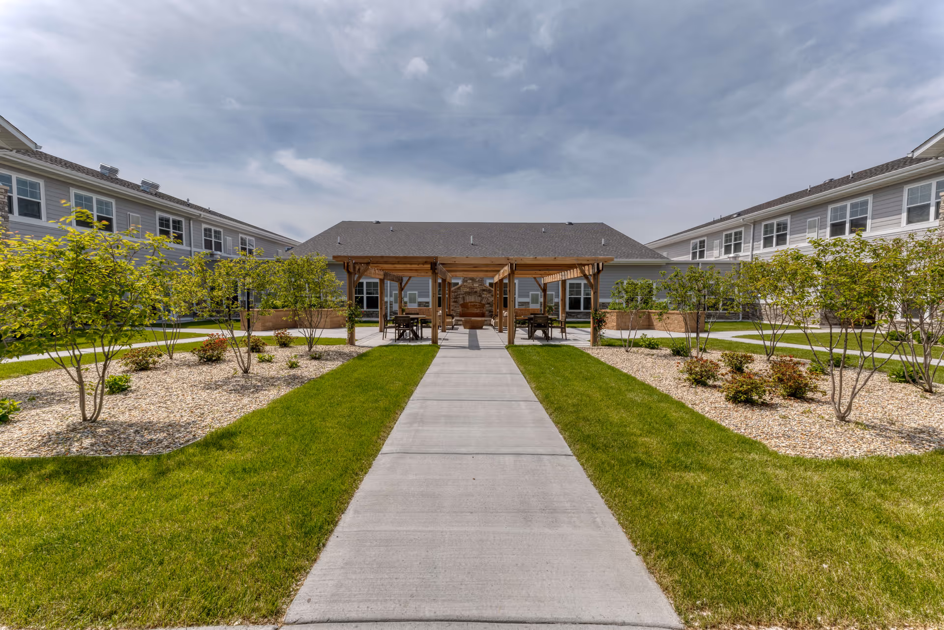 Outdoor courtyard area at Cedarhurst Senior Living of Dyer featuring a concrete walkway leading to a wooden pergola with seating and a stone fireplace. The courtyard is surrounded by green grass, small trees, and landscaped areas with rocks and shrubs. Two-story buildings with multiple windows flank the courtyard on both sides under a partly cloudy sky.