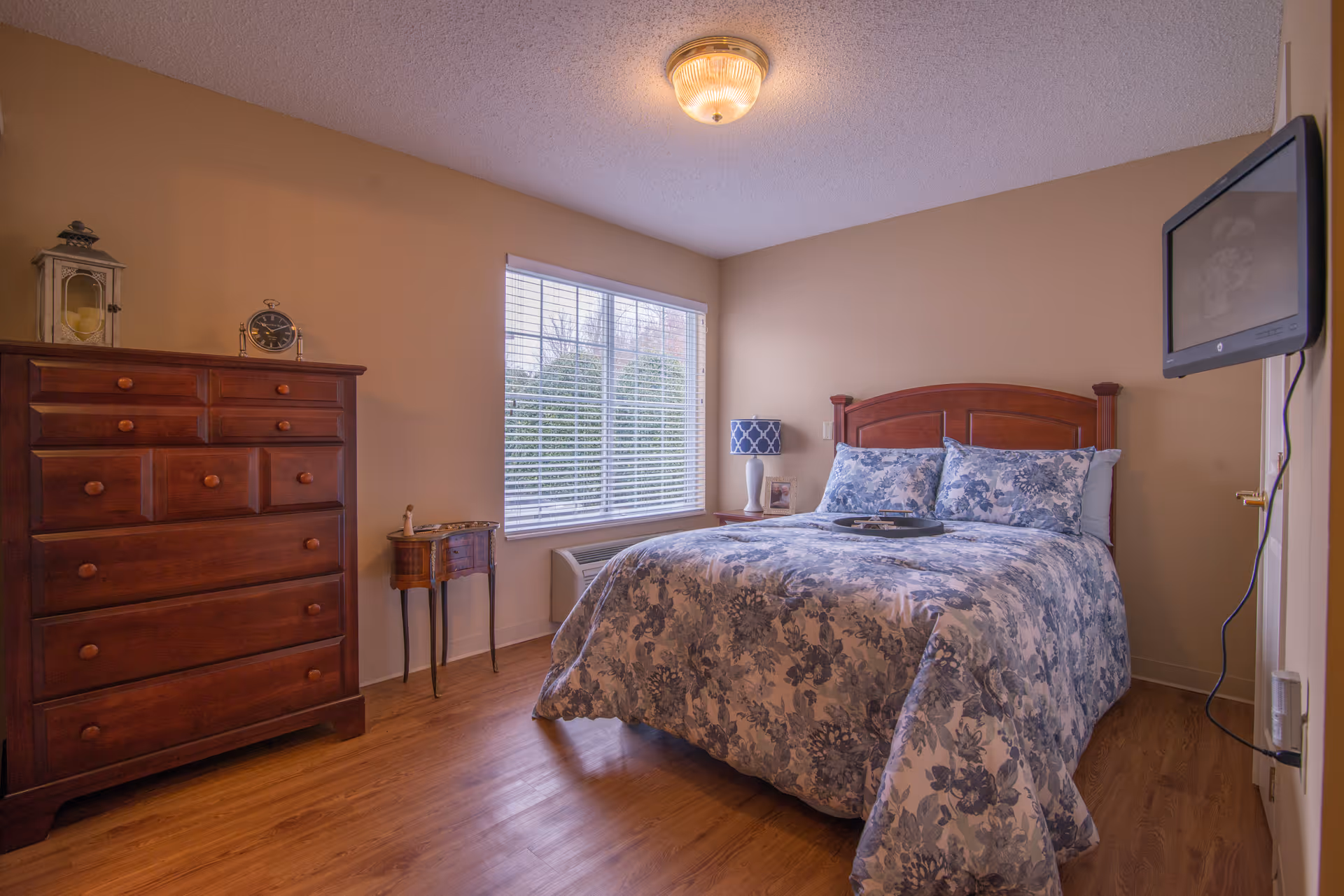 A cozy bedroom with a wooden bed frame and a bed made with blue and white floral bedding. There is a wooden dresser with a clock and a lantern on top, a small decorative table, and a window with white blinds letting in natural light. A flat-screen TV is mounted on the wall to the right.