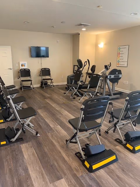 A fitness room with several black folding exercise chairs arranged in rows, each equipped with a yellow and black step platform. In the background, there are exercise machines including stationary bikes and elliptical trainers. A flat-screen TV is mounted on the beige wall, and a colorful motivational poster is displayed on the right wall. The room has wood-look flooring and recessed ceiling lights.