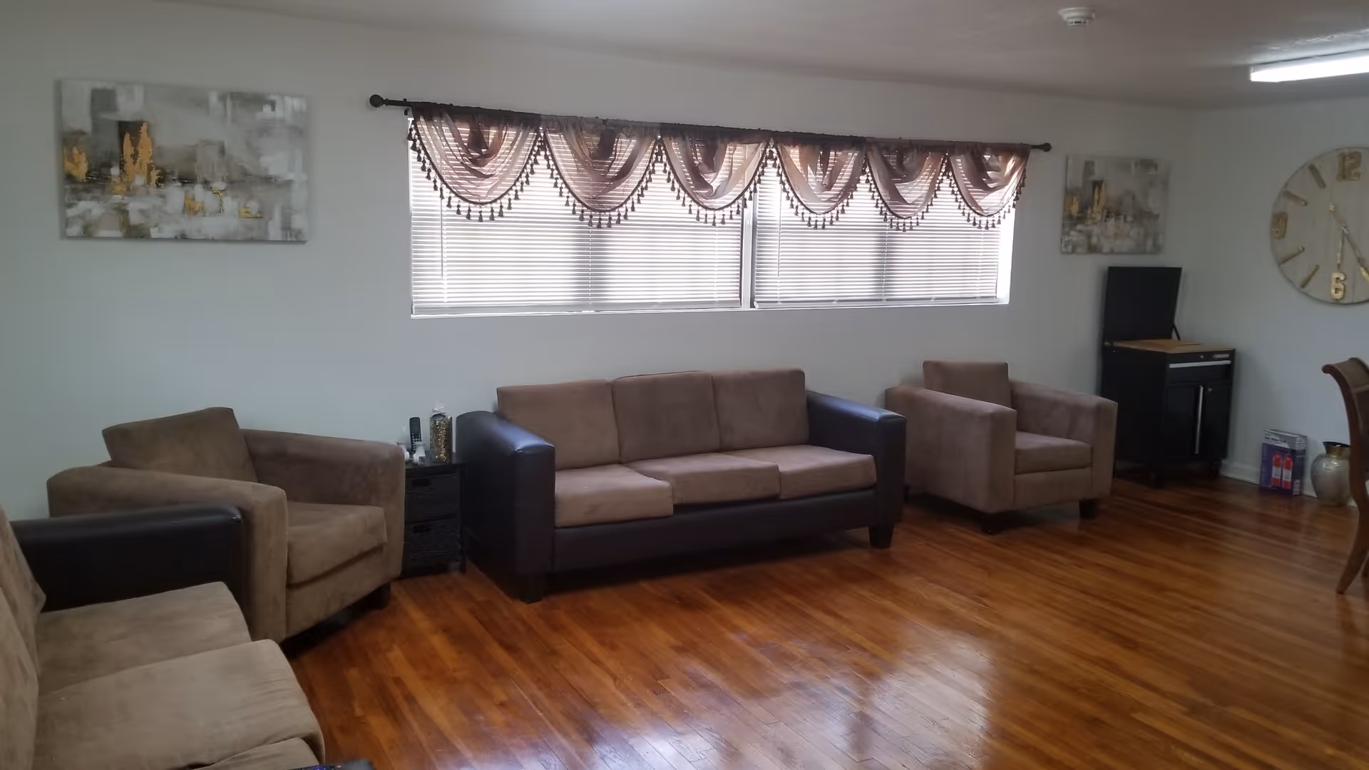 A living room with wooden flooring featuring a brown three-seater sofa and three matching armchairs arranged around the room. A window with blinds and decorative valances is centered on the wall behind the sofa. Two abstract paintings hang on the walls, and a large round clock is visible on the right side. A small black cabinet with a TV on top is in the corner.